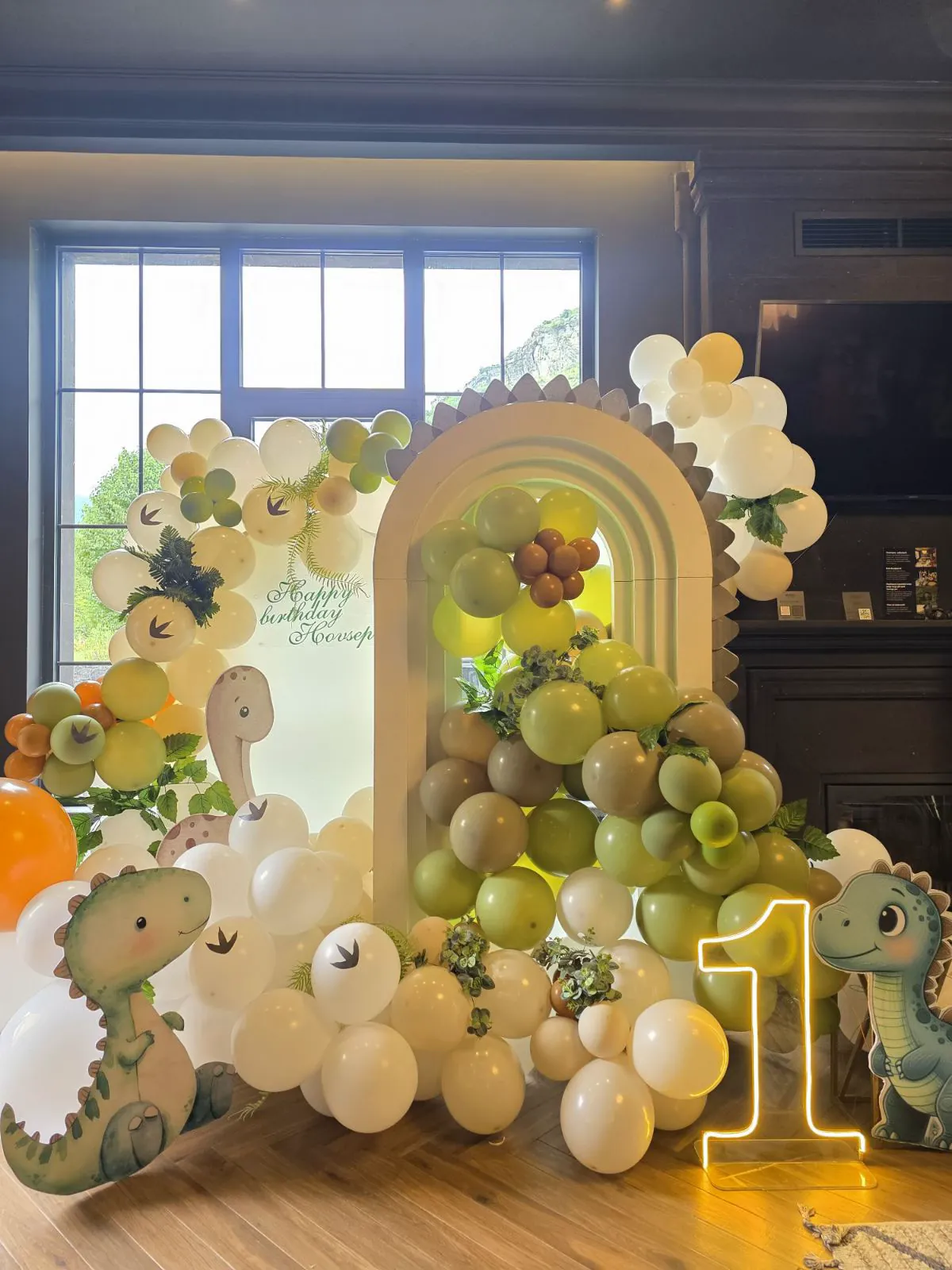 First-birthday decorations in the living room: green and white balloons, dinosaurs and a lit number '1'