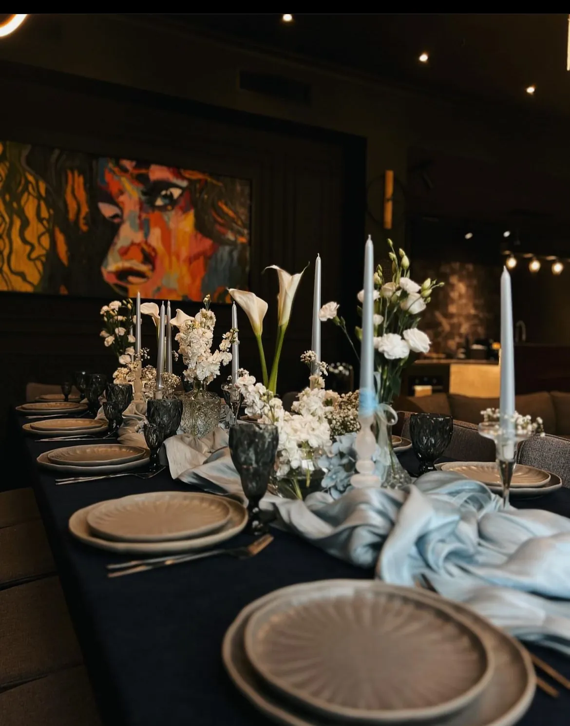 Table decorated with white flowers and pale blue candles, with the colourful painting behind